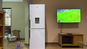 A kitchen with a white refrigerator, a wooden cabinet, a television displaying a soccer game, and a large metal bowl on the floor. - Oyibi New Furnished House - Charming 3 Bedroom (Accra)