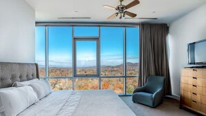 A modern hotel room with a large bed, a television, a chair, and a view of the landscape through large windows.