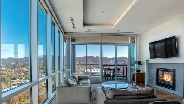 A modern living room with large windows, a fireplace, and a view of the city and mountains.