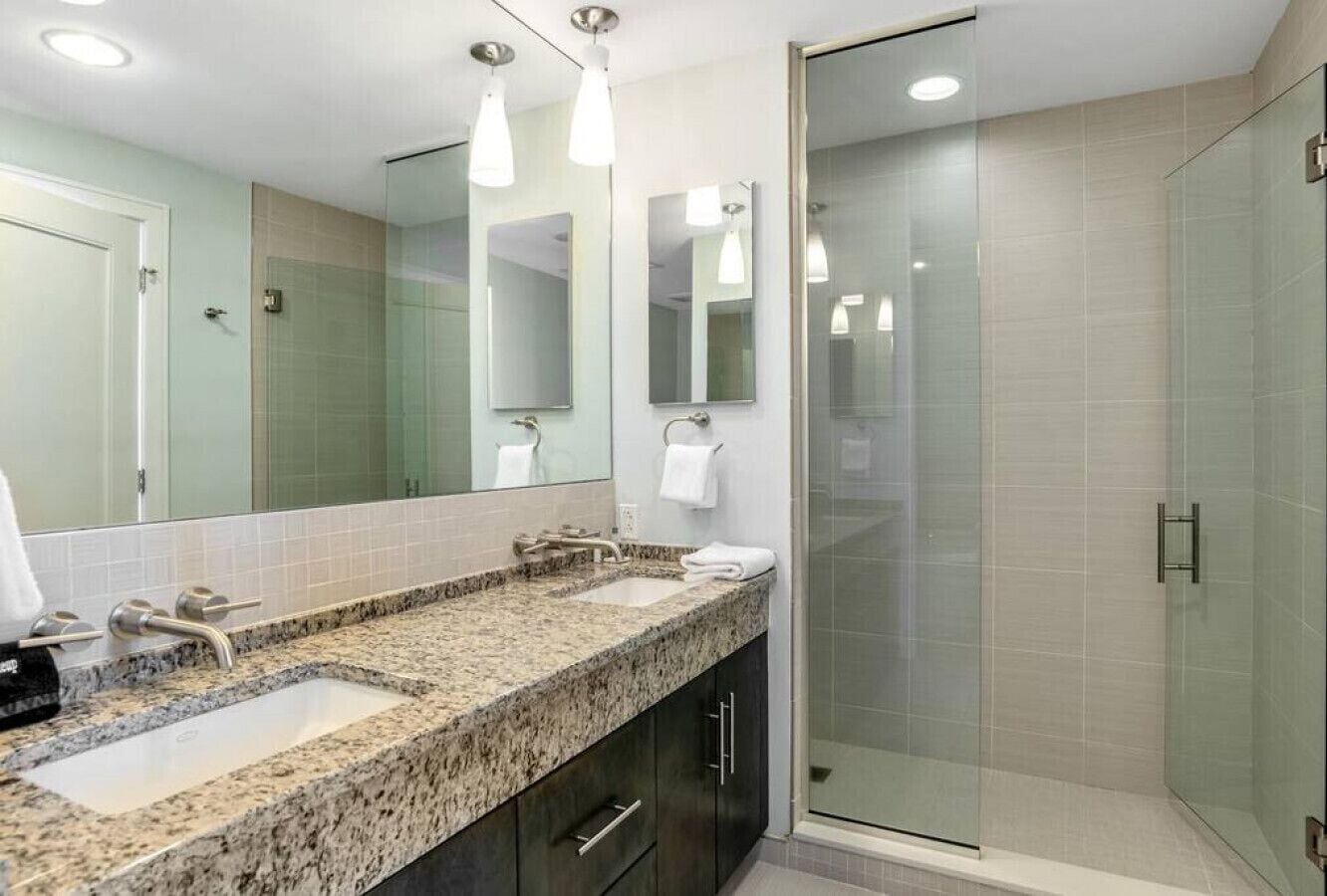 Une salle de bain moderne avec un comptoir en granit, deux lavabos, une douche en verre et un grand miroir.