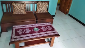 A room with a wooden sofa against a teal wall, a matching wooden table with a patterned tablecloth, and a tiled floor.