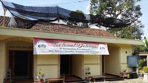 A building with a signboard reading "Selamat Datang di Homestay Punokawan" and contact information. There are potted plants and a bench outside.