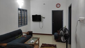 A living room with a sofa, a television mounted on the wall, a fan, a clock, and a door. - wellmark resort room 1, Hall 1,  bathroom 1 (jambavanodai)
