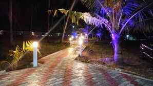 A paved pathway with patterned tiles leading to a well-lit area with palm trees and street lamps. - wellmark resort room 1, Hall 1,  bathroom 1 (jambavanodai)