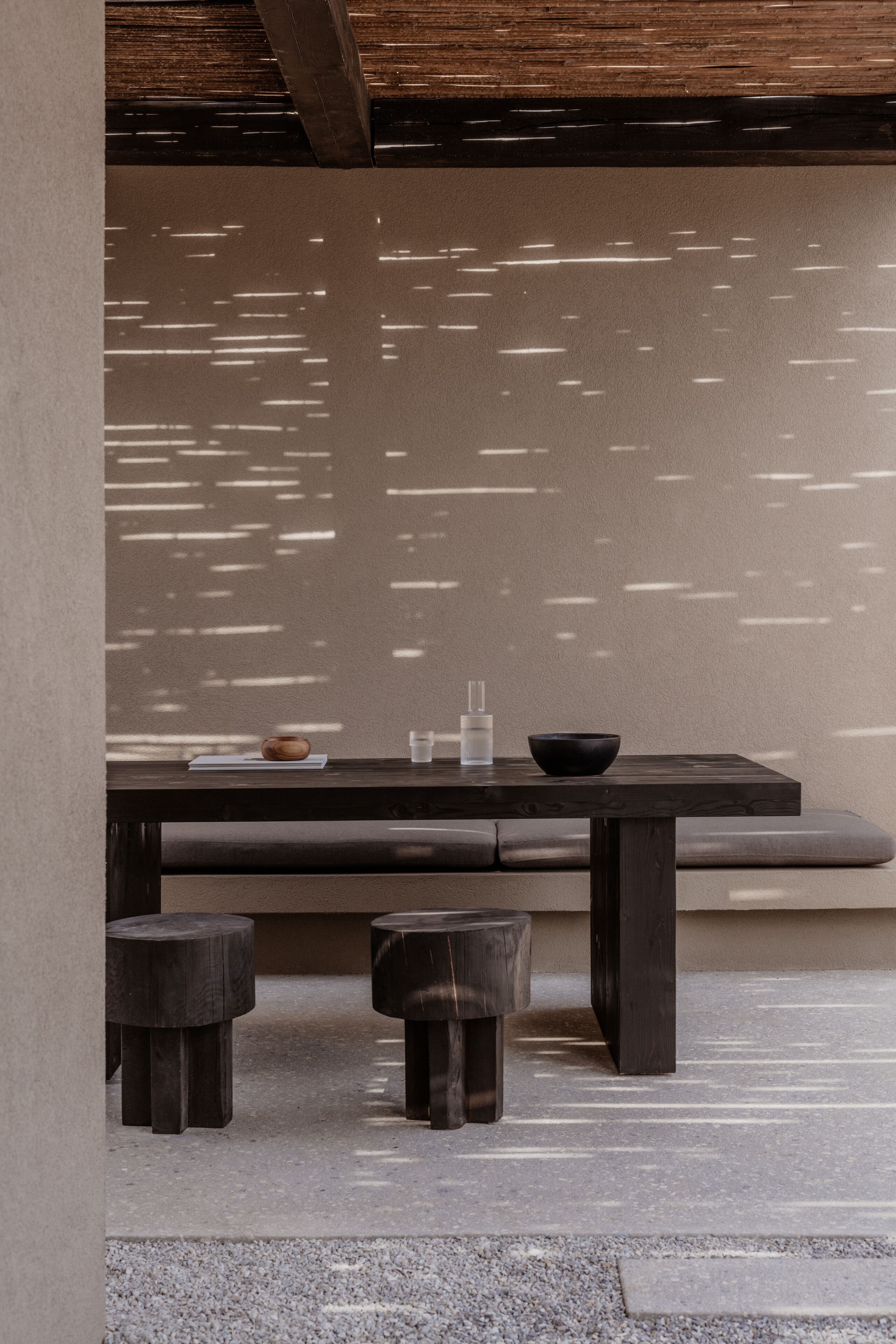 A minimalist interior with a wooden bench, two stools, and a table with a bowl and glasses.