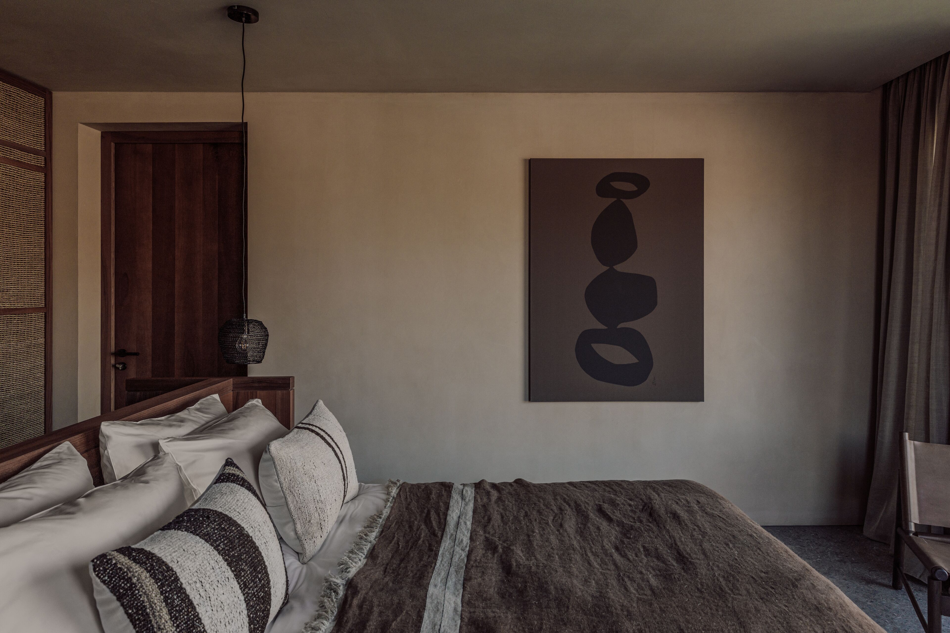 Un dormitorio con cama, almohadas, una colcha, una obra de arte en la pared, una puerta de madera y una silla.