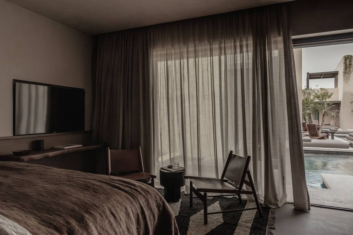 A modern hotel room with a large bed, a flat-screen TV, a desk, and a chair. The room has a large window with sheer curtains and a view of a pool and outdoor seating.