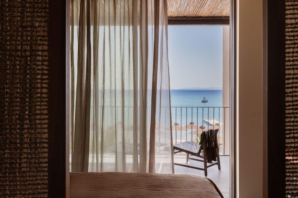 A room with a view of the sea through sheer curtains, a wooden bed, and a balcony with a chair and a table.