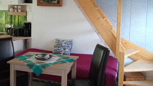 A dining area with a wooden table, chairs, and a red sofa. A staircase with wooden railings is visible in the background.