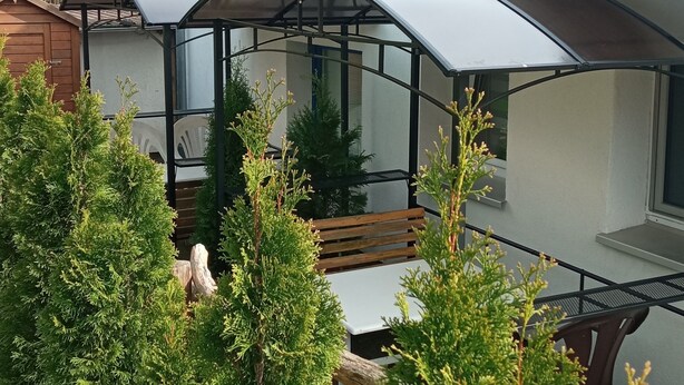 Una casa moderna con tetto rosso, un'area esterna coperta con una panchina e vegetazione circostante.