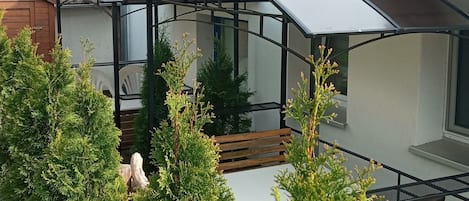 A modern house with a red roof, a covered outdoor area with a bench, and surrounding greenery.