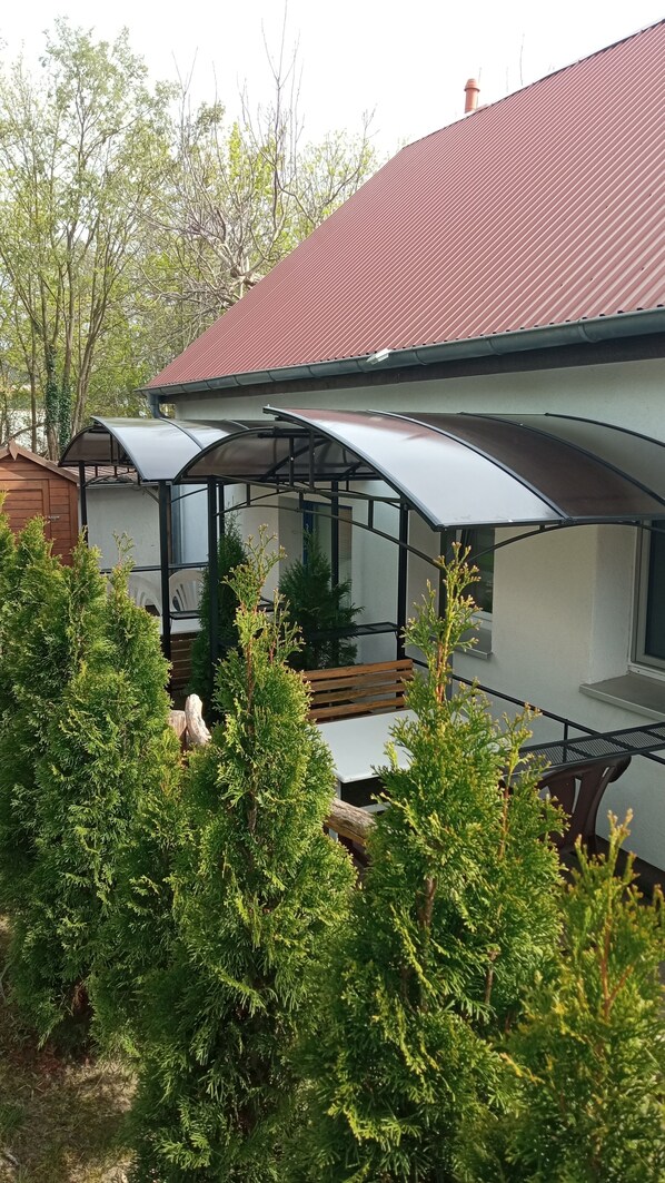 Une maison moderne avec un toit rouge, un espace extérieur abrité doté d’un banc, et une végétation luxuriante tout autour.