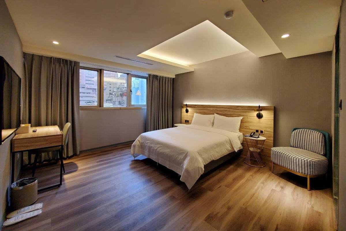 A modern hotel room with a large bed, a desk, a chair, and a view of the city through the window.