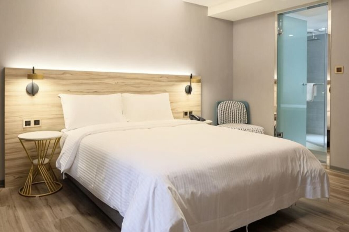 A modern hotel room with a large bed, bedside tables, a chair, and a glass shower door.