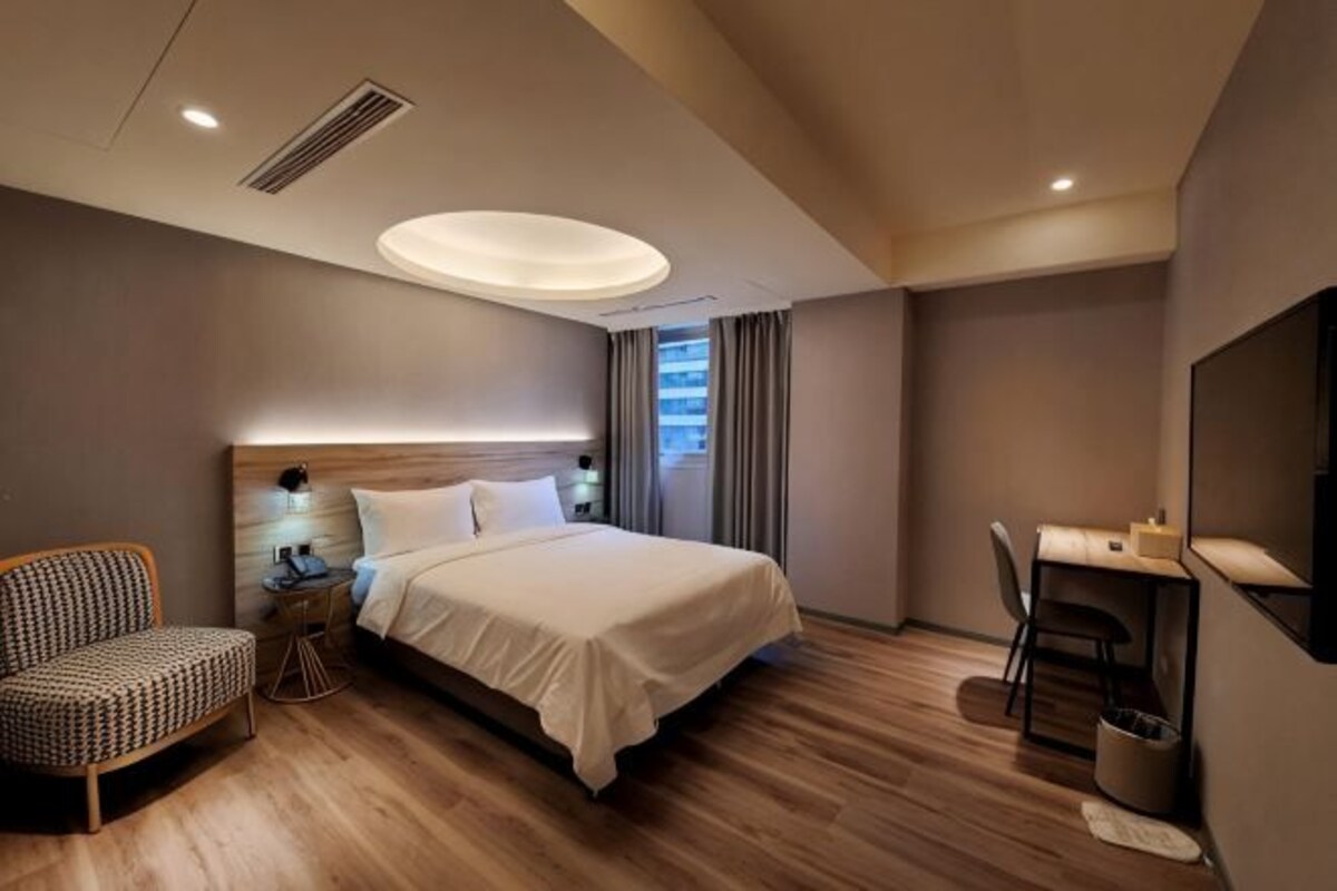 A modern hotel room with a large bed, a desk, a chair, and a bedside table.