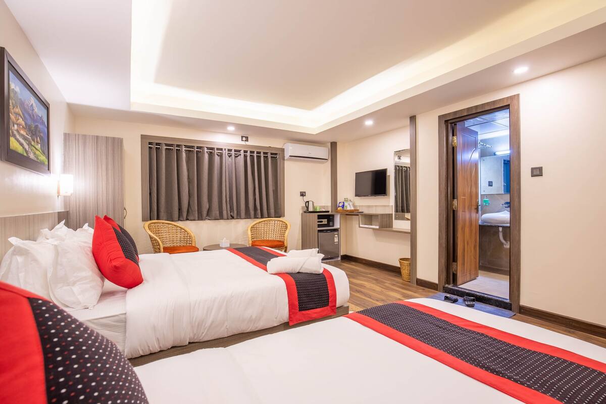 A hotel room with two beds, a television, a desk with a computer, and a bathroom visible through a glass door.