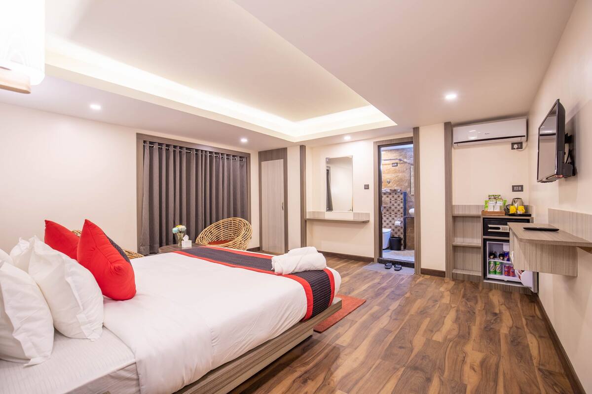 A modern hotel room with a large bed, a wooden floor, a TV mounted on the wall, and a small kitchenette area.