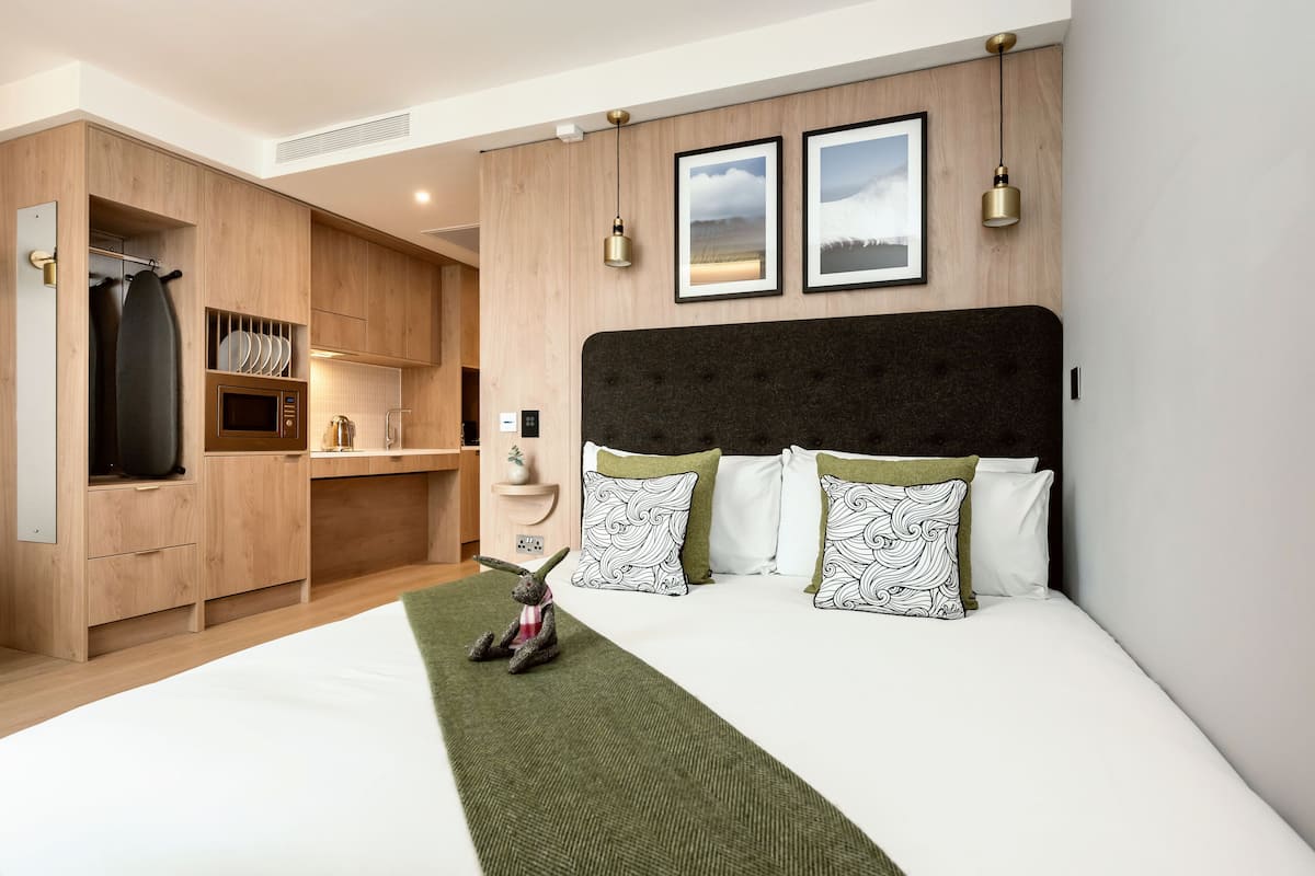 A modern hotel room with a large bed, a wooden wardrobe, a kitchenette, and a view of the outdoors.