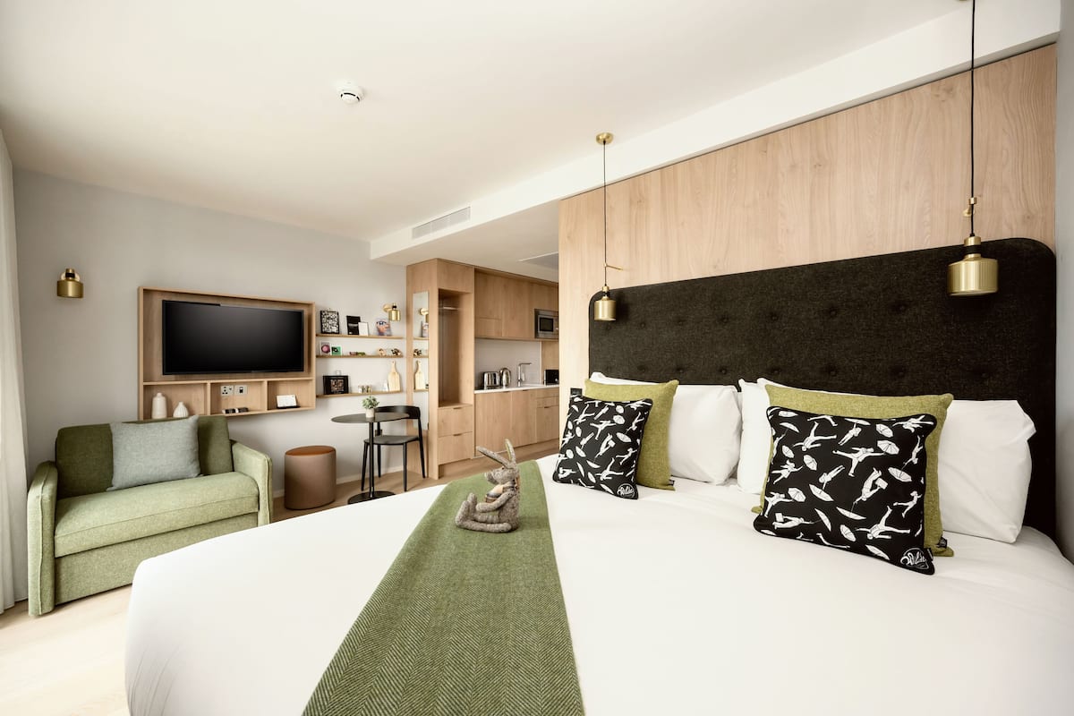 A modern hotel room with a large bed, a TV mounted on the wall, a kitchenette, and a seating area with a sofa and chairs.