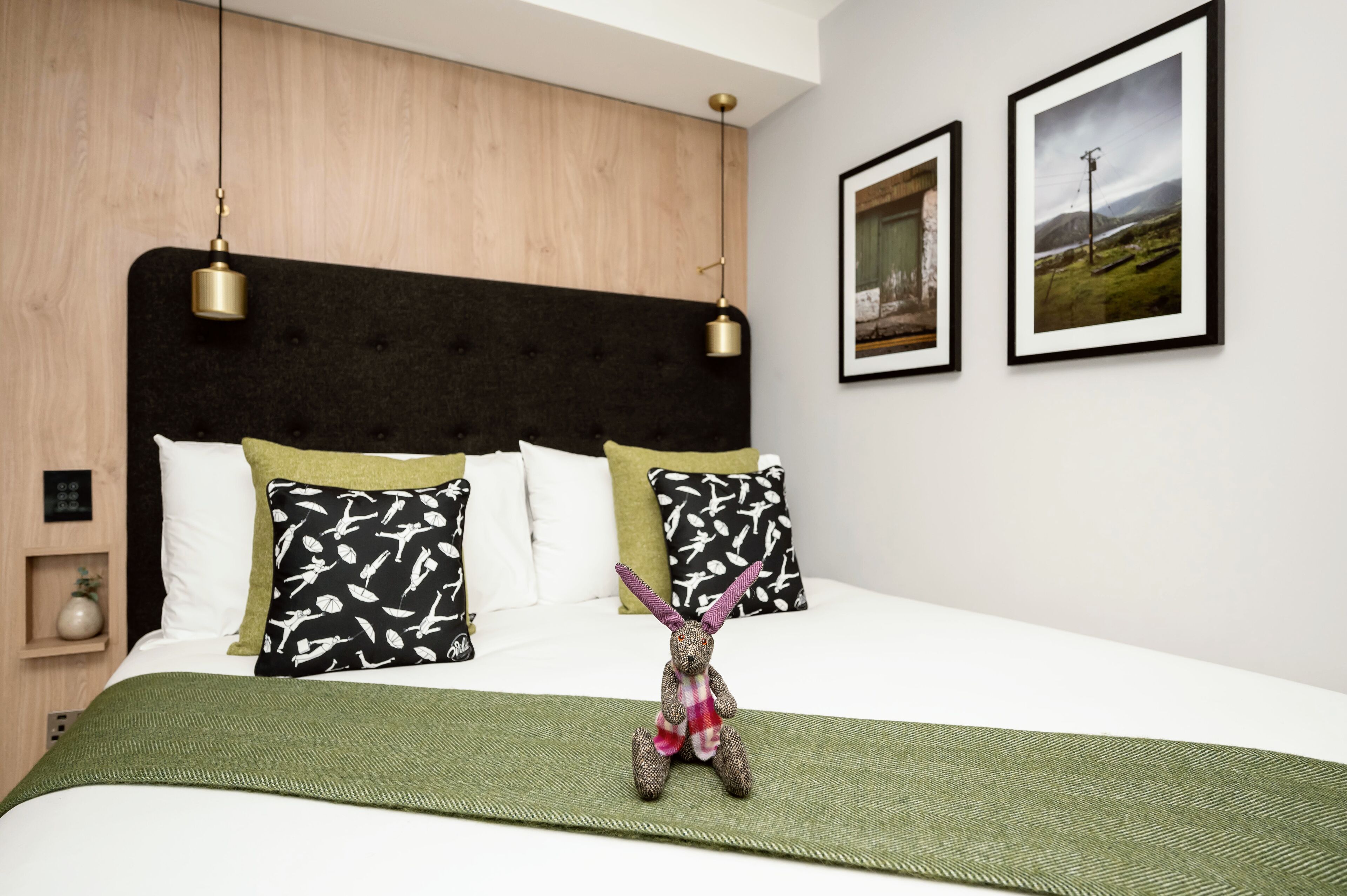 Una cama bien hecha con una colcha verde, dos almohadas de estampado blanco y negro, y un pequeño peluche sobre la cama.