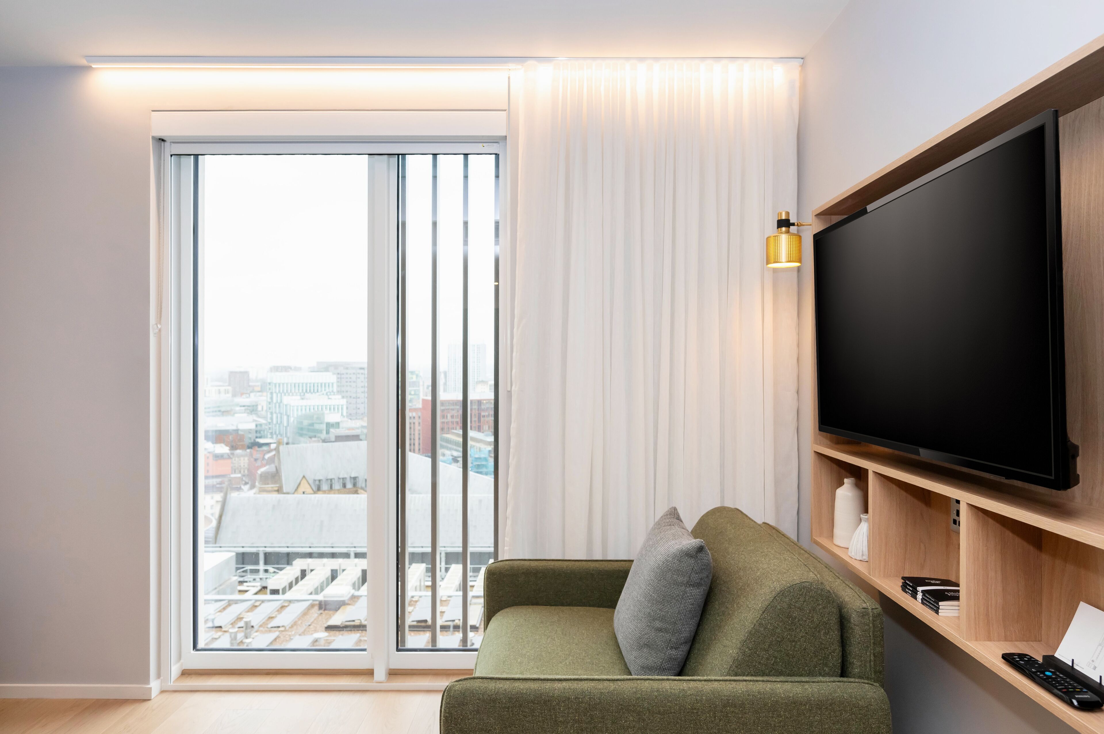 A modern living room with a green sofa, a large flat-screen TV, and a sliding glass door leading to a city view.