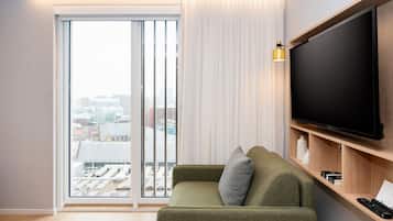 A modern living room with a green sofa, a large flat-screen TV, and a sliding glass door leading to a city view.
