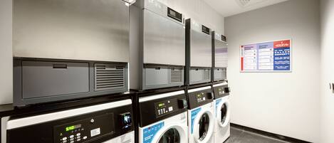 A row of front-loading washing machines in a commercial laundry room with a control panel and a posted laundry schedule.