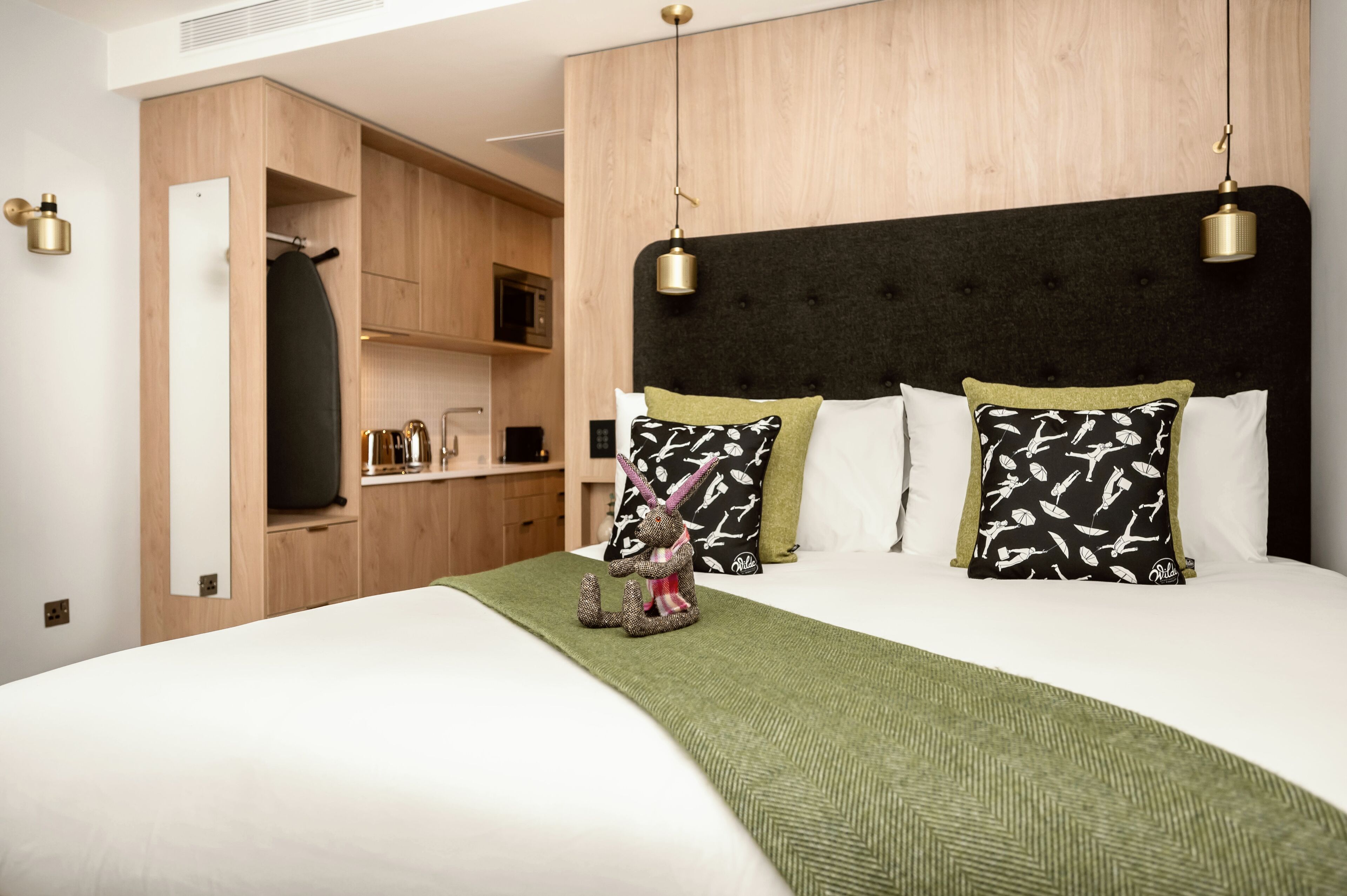 Una cama impecable con un cubrecolcha verde, cabecera con dos lámparas colgantes y una pequeña figura decorativa de conejo.