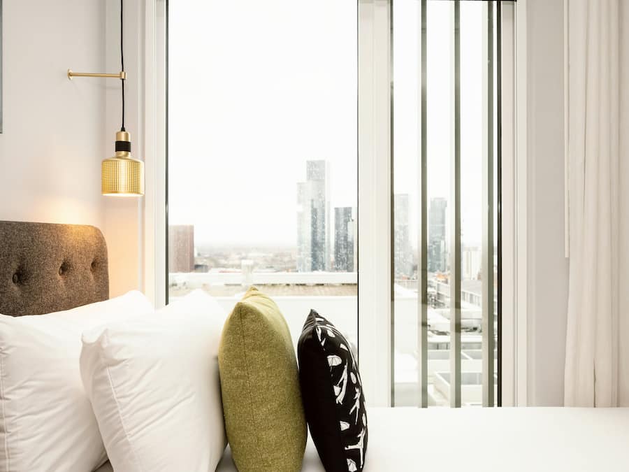 A modern hotel room with a city view, a tufted headboard, and a hanging light fixture.