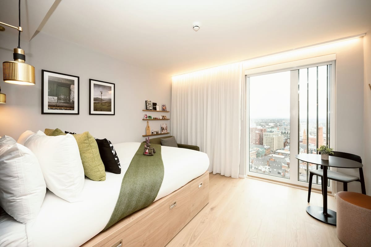 A modern bedroom with a large bed, a city view through the window, and a small table with a chair.