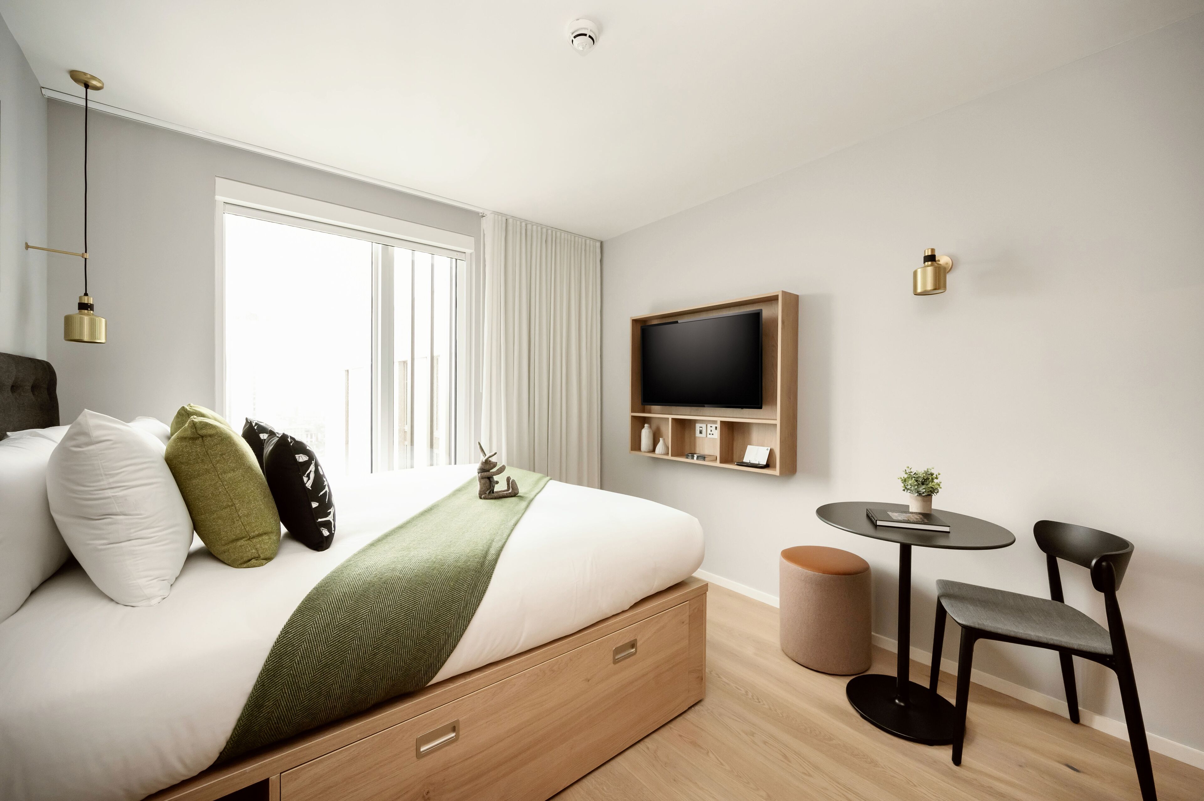 A modern hotel room with a bed, bedside table, chair, and a TV mounted on the wall.