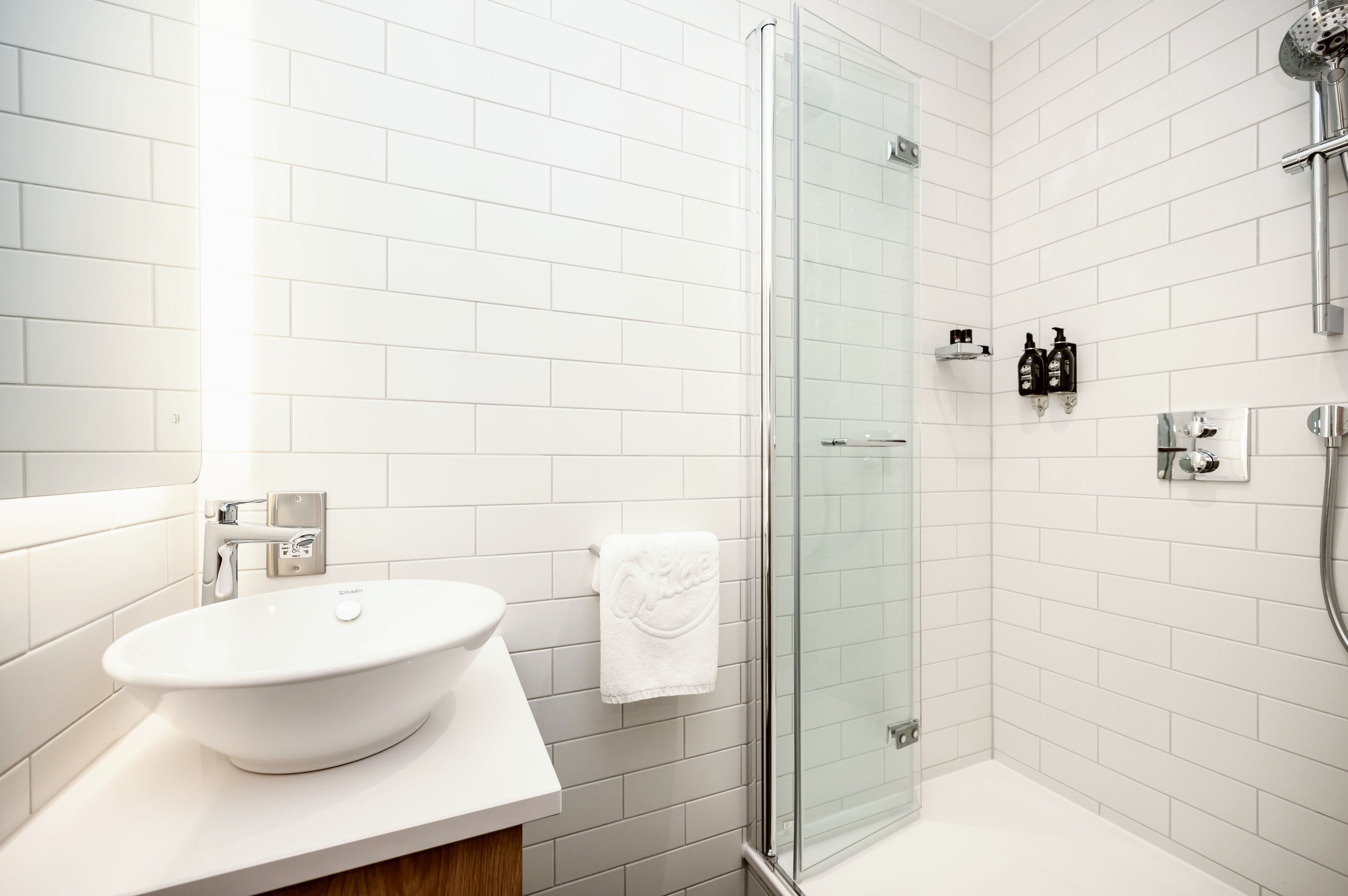 Un bany modern amb un lavabo blanc, un plat de dutxa amb envàs de vidre i una paret revestida de rajoles blanques.
