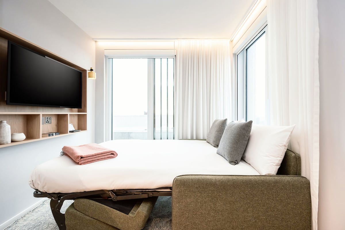 A hotel room with a bed, a flat-screen TV mounted on the wall, a bedside table with a lamp, and a view of the outdoors through large windows.