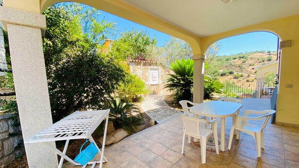A covered patio with a table and chairs, a white metal chair with a blue bag, and a stone wall. - Sa Domo Lada 2 (San Teodoro)