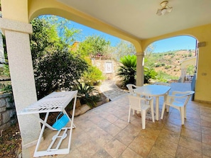 A covered patio with a table and chairs, a white metal chair with a blue bag, and a stone wall.