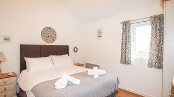 Una cama bien hecha con una colcha gris estampada, un cabecero de madera y dos toallas blancas enrolladas.