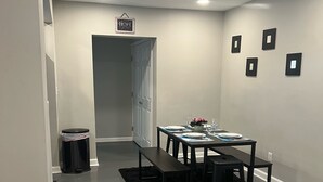 Uma sala de jantar com uma mesa posta para quatro pessoas, uma lixeira e uma porta que dá para outro quarto.