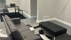 Uma sala de estar moderna com um sofá cinza, uma mesa de centro preta e um banquinho preto. Há uma pequena mesa com uma cesta de objetos ao fundo.