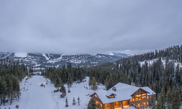 Eine Lodge mit schneebedecktem Dach, umgeben von Wald und Bergen im Hintergrund.