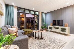 A modern living room with a grey sofa, a coffee table, a TV on a white stand, and a large window with curtains.