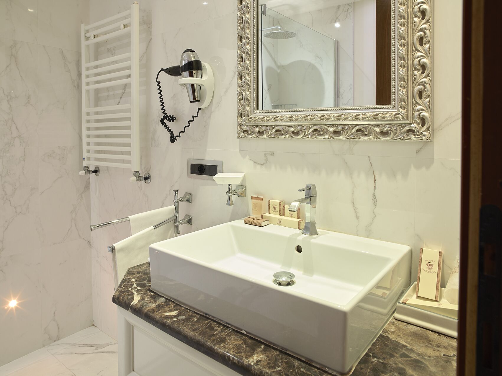 Une salle de bain avec un grand lavabo blanc, un miroir doté d’un cadre orné, un porte-serviettes et un sèche-cheveux portatif fixé au mur.