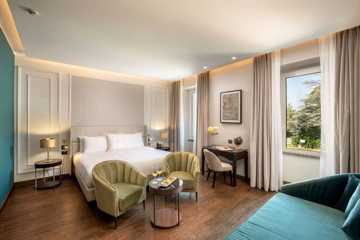 Une chambre d’hôtel avec un grand lit, deux fauteuils, une petite table avec un verre, et une vue sur la verdure à l’extérieur.