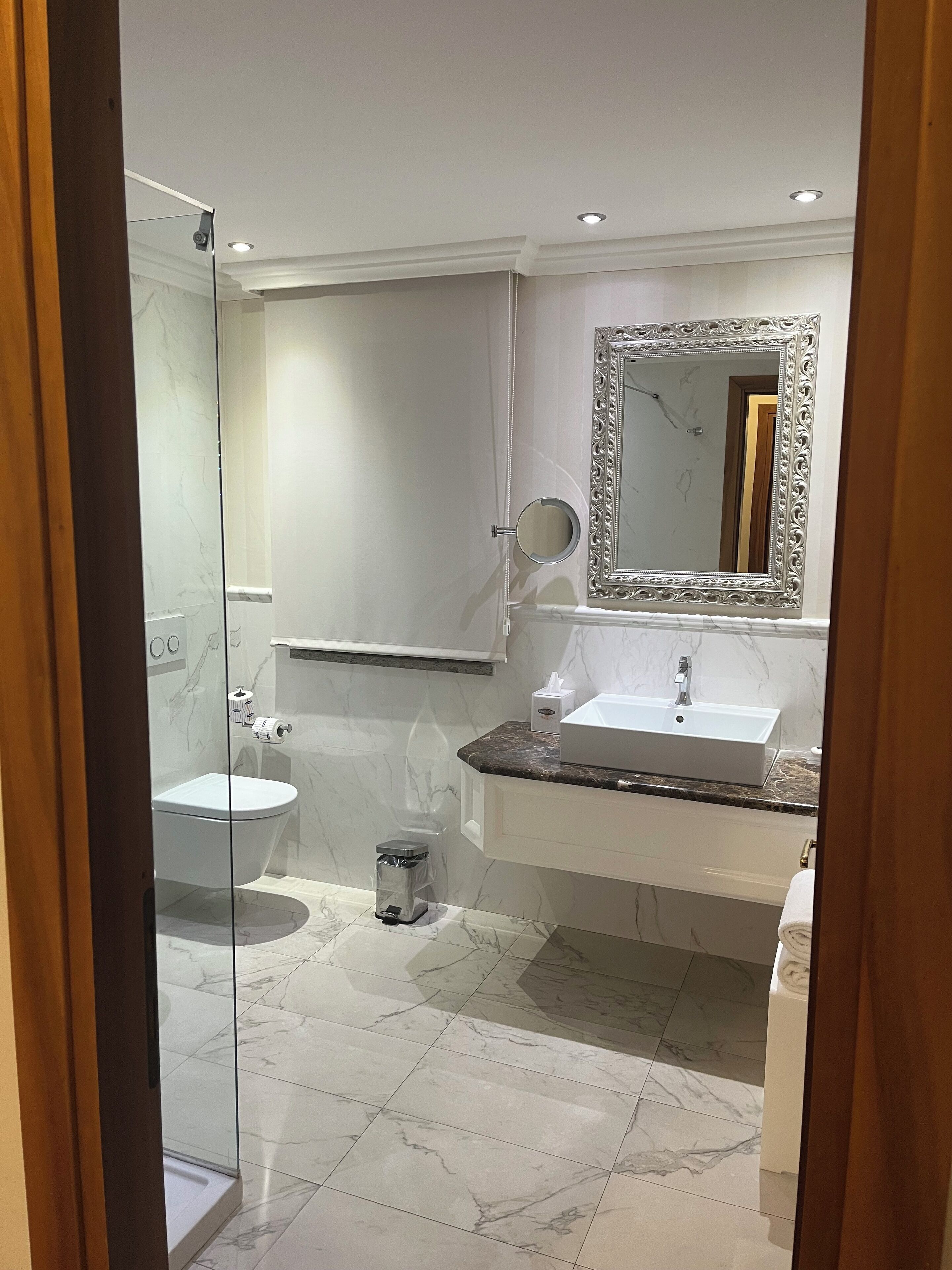 Et moderne badeværelse med en glas brusekabine, et hvidt toilet, en vask med marmorbordplade og et stort spejl med en detaljeret ramme.