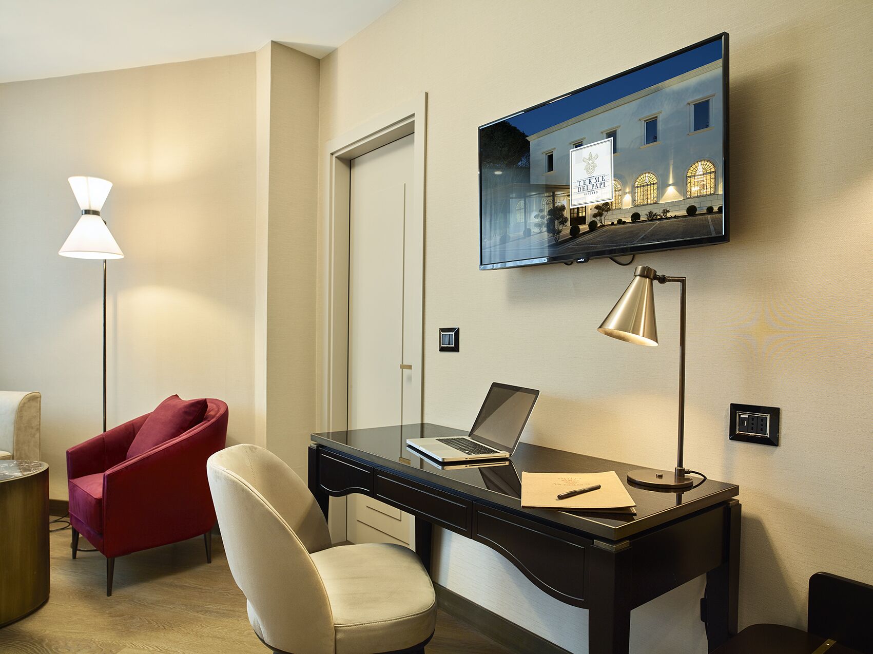 Une chambre d’hôtel équipée d’un bureau, d’une chaise rouge, d’une chaise blanche, d’un ordinateur portable, d’un téléviseur fixé au mur et d’un lampadaire.