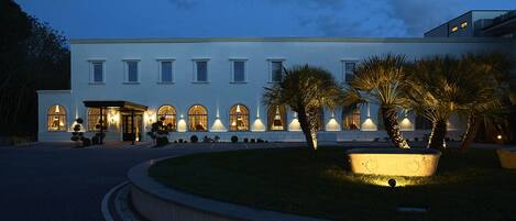 Un hotel moderno con fachada simétrica, ventanas en arco y un jardín ajardinado con palmeras.