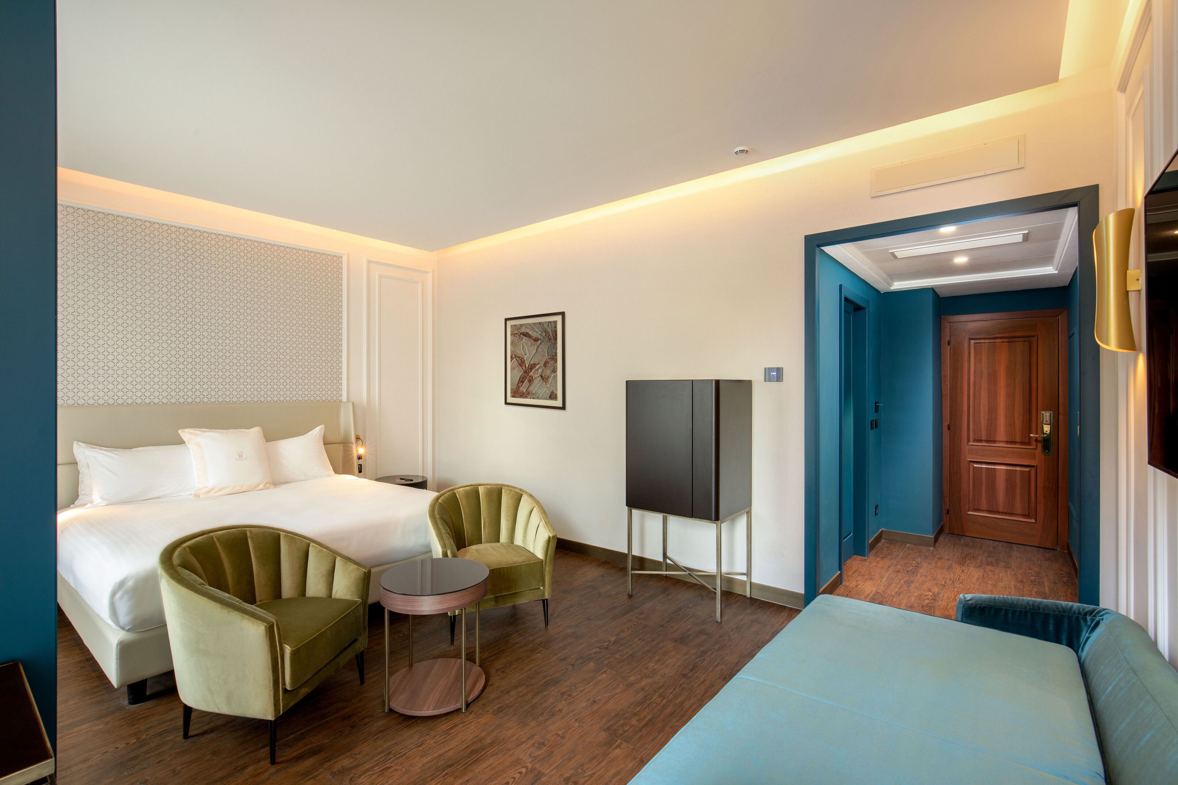 Ein Hotelzimmer mit einem Bett, zwei Sesseln, einem kleinen Tisch, einem Fernseher und einer Tür zu einem anderen Raum.