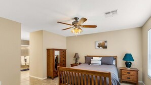 Un dormitorio con cama, mesita de noche, cómoda, ventilador de techo, rejilla de ventilación en la pared y ventana con persianas.