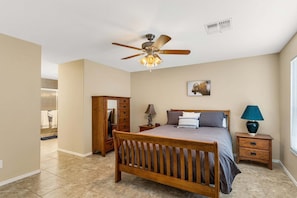 A bedroom with a bed, a nightstand, a dresser, a ceiling fan, a wall-mounted air vent, and a window with blinds.