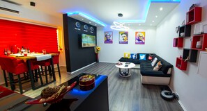 Moderni olohuone, jossa on ruokailutila. Huoneen sisustukseen kuuluu musta sohva, punaiset tuolit, sohvapöytä ja seinälle asennettu televisio.