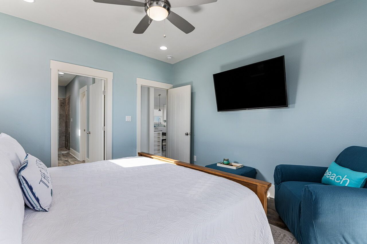 A bedroom with a bed, a ceiling fan, a TV, and a door leading to a bathroom.